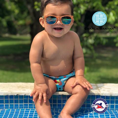 Baby in sunglassess sitting on the side of a pool wearing a Charlie Banana Swim Diaper